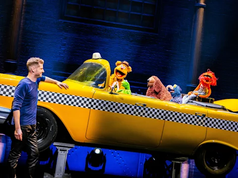 A man stands beside a yellow taxi on stage with several Muppet characters sitting inside under blue stage lighting.