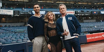 Three people stand on a baseball field near home plate. The stands are mostly empty. One person is holding a baseball. All three wear navy or black clothing.