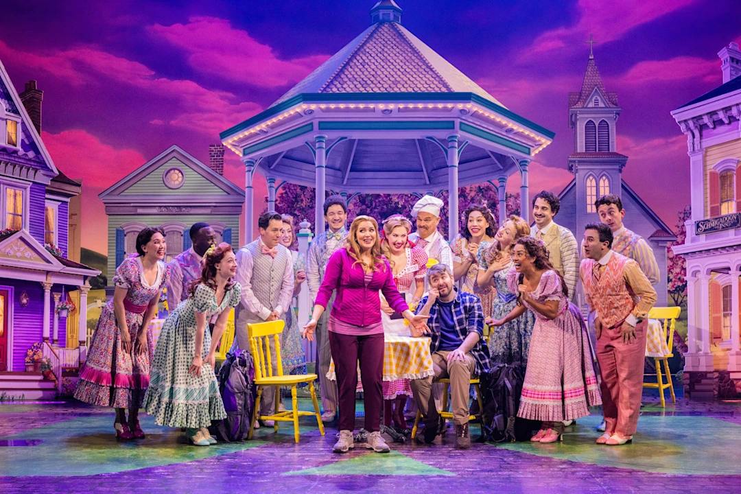 A theater cast in colorful costumes gathers around a woman holding a pie, standing in front of a gazebo on a brightly lit, whimsical stage set resembling a cheerful town.