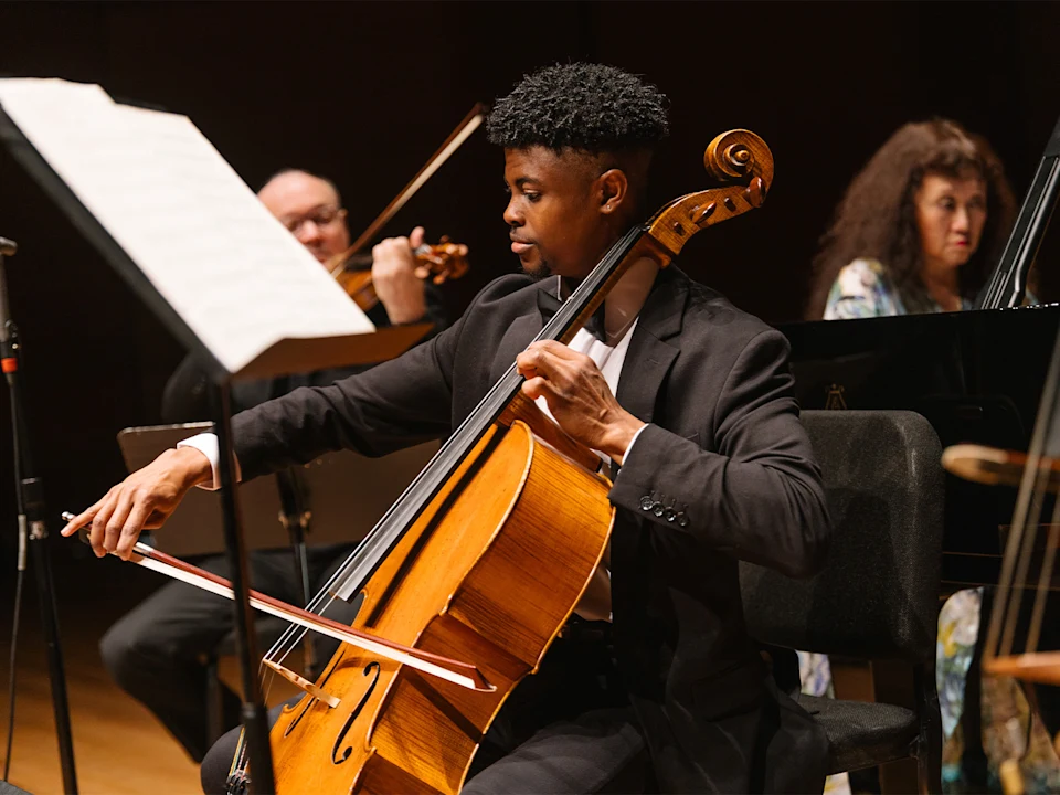 A cellist in a suit plays his instrument with a bow during an orchestral performance, with other musicians visible in the background.