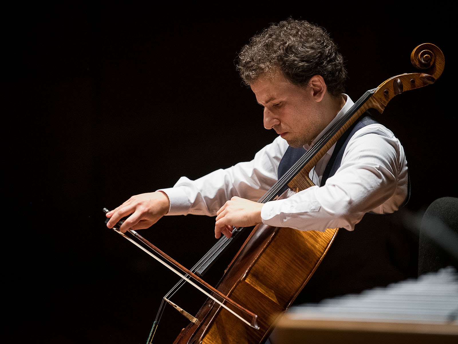 Production photo of Chamber Music Society of Lincoln Center: Summer Evenings II in New York, showing a man wearing a white shirt and vest plays a cello with a bow on stage, focusing intently on the instrument.
