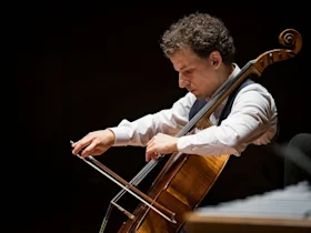 Production photo of Chamber Music Society of Lincoln Center: Summer Evenings II in New York, showing a man wearing a white shirt and vest plays a cello with a bow on stage, focusing intently on the instrument.