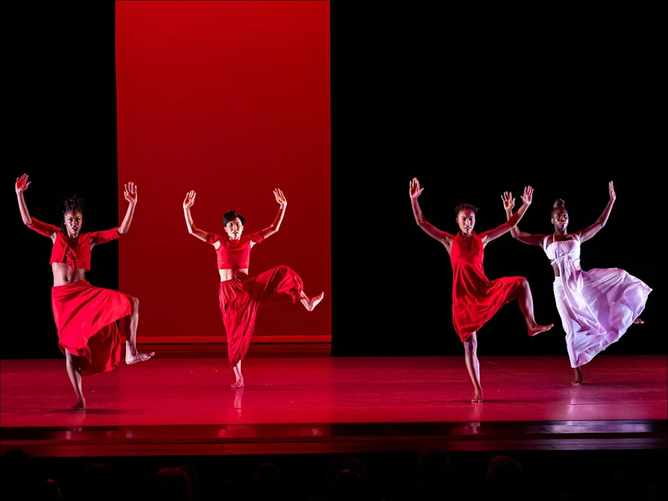Four dancers perform on stage against a red and black backdrop. Three wear red outfits, one wears white, and all have one leg lifted and both arms raised.