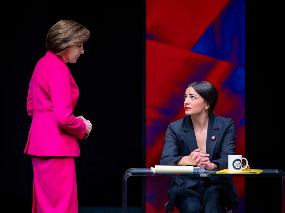 Two women in business attire converse; one standing in a bright pink suit, the other sitting at a desk in a black suit, appearing attentive. A red and blue background accentuates the scene.