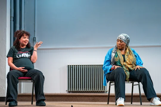 Two performers sit on chairs onstage; a pregnant woman in a black T-shirt gestures while an older woman in a blue jacket and headscarf looks on.