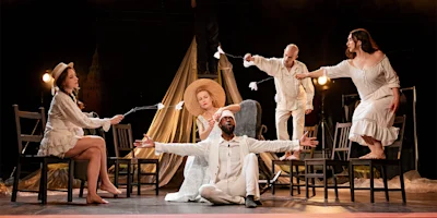 Five actors in white costumes perform on stage with chairs, sticks with white tips, and theatrical props, creating a dramatic and expressive composition.
