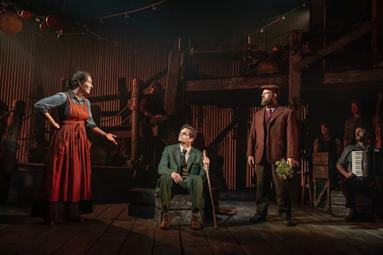 Actors perform on a rustic stage, including a woman in a red apron, a man sitting with a cane, a man in a brown suit holding flowers, and a person playing an accordion.