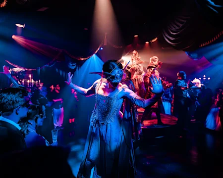 People in elaborate costumes and masks dance under dramatic lighting at a masquerade ball, with onlookers gathered around them in a dimly lit, decorated venue.