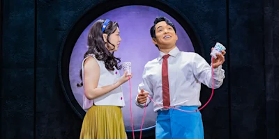 Two actors on stage use a string telephone, standing in front of a circular window; one holds a paper cup, the other holds a model of teeth.