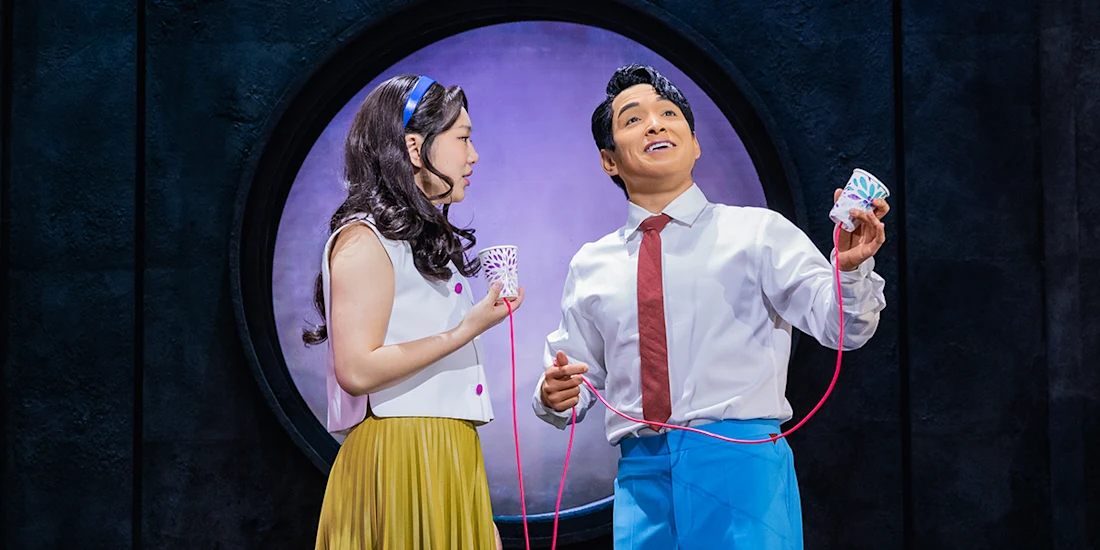 Two actors on stage use a string telephone, standing in front of a circular window; one holds a paper cup, the other holds a model of teeth.