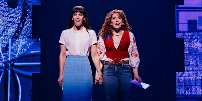 Two women stand on stage holding hands. One wears a white blouse and blue skirt, the other a red vest and embroidered blouse with jeans. Both look forward under stage lighting.