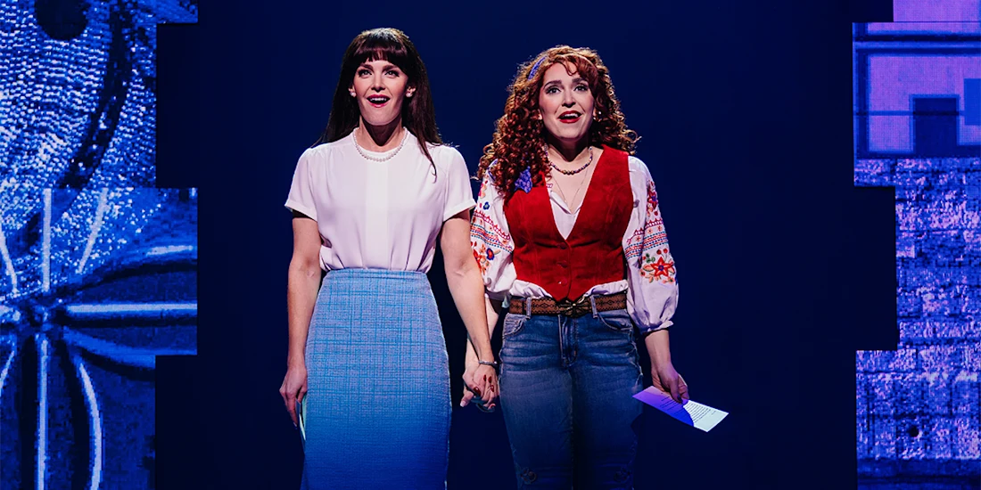 Two women stand on stage holding hands. One wears a white blouse and blue skirt, the other a red vest and embroidered blouse with jeans. Both look forward under stage lighting.