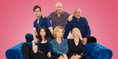 Six people posed together on a blue couch against a solid pink background, with three sitting in front and three standing behind them.