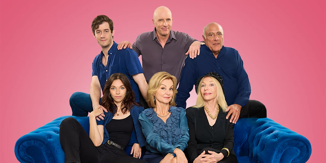 Six people posed together on a blue couch against a solid pink background, with three sitting in front and three standing behind them.