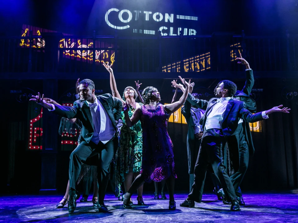 A group of dancers, dressed in 1920s attire, perform energetically under neon signs reading "Cotton Club" on a dimly lit stage.