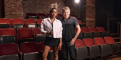 Two people stand in front of empty theater seats, posing for a photo; one is dressed in a white jacket and skirt, the other in a black shirt and pants.
