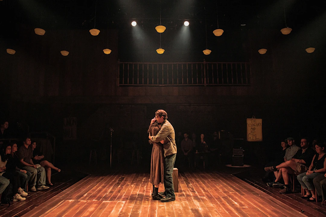 Two actors embrace in the center of a dimly lit stage, surrounded by an audience seated on wooden benches.
