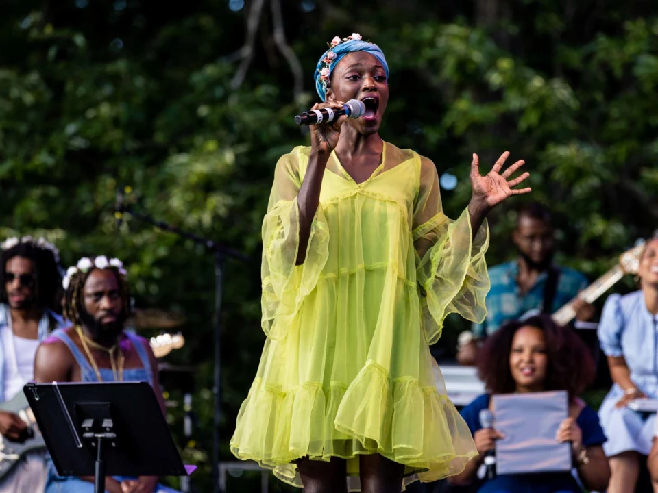 A person in a bright yellow dress sings into a microphone on an outdoor stage, with band members and backup singers visible in the background.