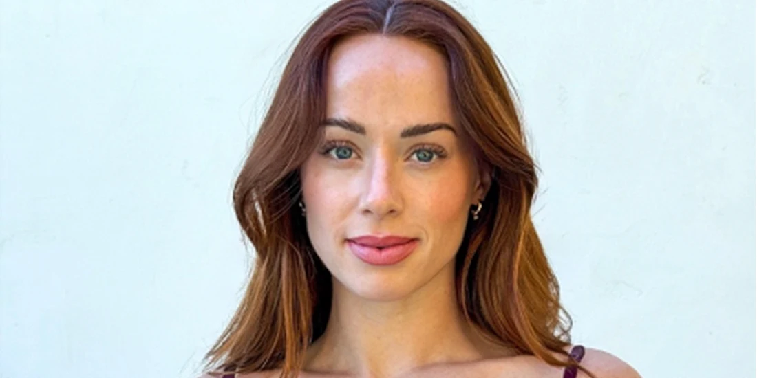 A woman with long auburn hair and light eyes stands in front of a plain white background, looking directly at the camera.