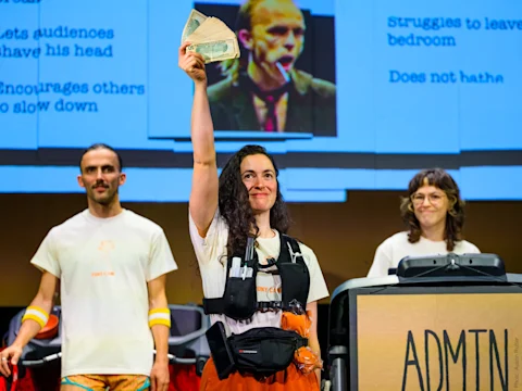 A woman stands on stage holding cash aloft, flanked by two people, with a screen displaying text and an image in the background.