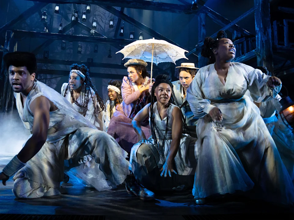 A cast of seven performers in period-style clothing perform a scene on a dimly lit wooden stage, with one central figure holding a white lace parasol.