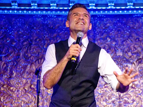 A man in a white shirt and dark vest holds a microphone and smiles while performing on stage with ornate blue and gold background decor.