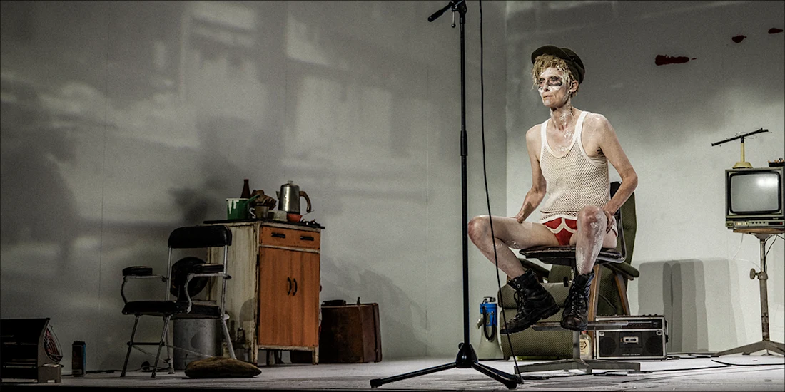 A performer in a white tank top and red underwear sits on a chair on a stage with a microphone, surrounded by a cabinet, a television, and a suitcase.