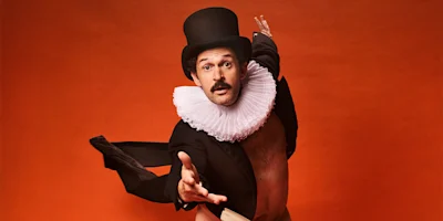 A man in a top hat, ruffled collar, and tuxedo jacket poses dramatically with one arm extended against an orange background.