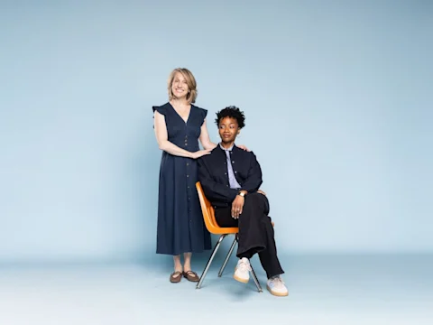 A standing woman rests her hand on the shoulder of a seated woman against a plain light blue background.