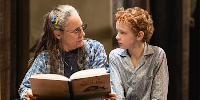 An older woman with glasses reads a book to a young boy with curly red hair; both are wearing patterned shirts and sitting closely together.