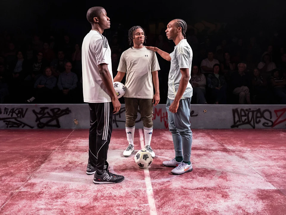 Three young men stand on a red sports court with soccer balls at their feet, surrounded by graffiti walls and an audience watching in the background.