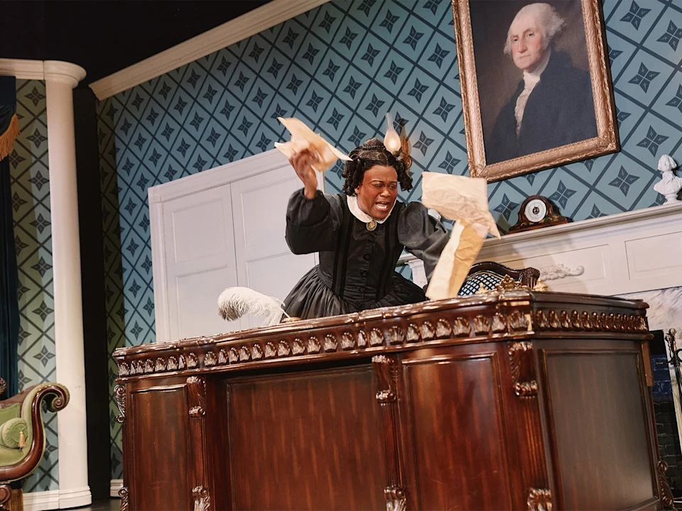 A wax figure of a woman in 19th-century attire angrily crumples papers at a large wooden desk in an ornate room, with a portrait of George Washington on the wall.