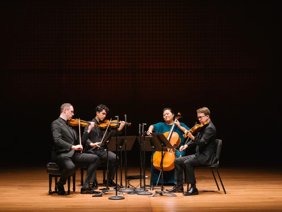 Four musicians in formal attire perform a string quartet on a stage. They are seated closely, each playing a different string instrument: two violins, a viola, and a cello.