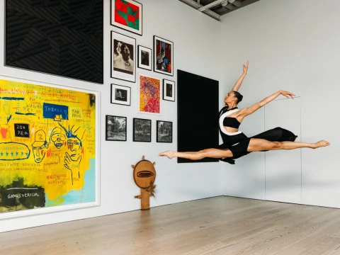 A dancer performs a mid-air leap in an art gallery, surrounded by various framed artworks and a large painting with vibrant colors.