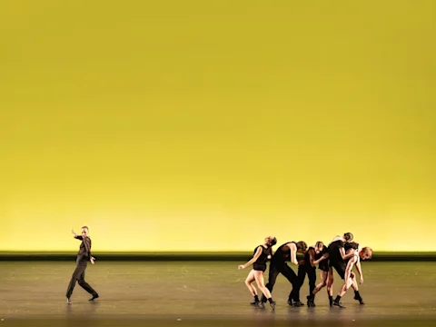 Dancers perform on stage with a yellow background; one dancer is separate from a group on the right.