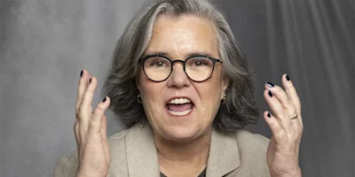 A person with gray hair and glasses, wearing a beige blazer, gestures with raised hands while speaking against a gray background.