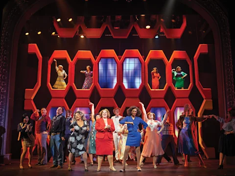 A large cast performs on stage in colorful costumes, with some actors standing in illuminated hexagonal set pieces, under theatrical lighting.