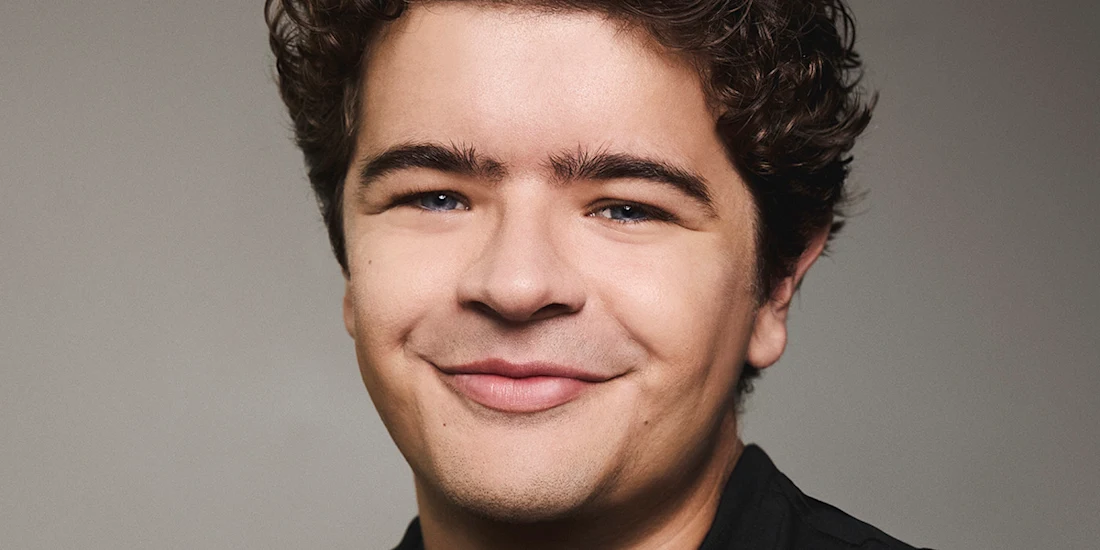 A young man with curly brown hair, fair skin, and blue eyes smiles at the camera in front of a plain gray background.