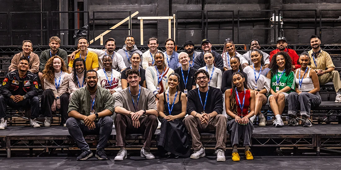 A large group of people pose together on tiered platforms in a theater setting, all wearing lanyards and casual clothing.