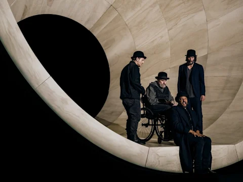 Production photo of Waiting for Godot on Broadway with the cast in dark clothing and bowler hats pose on a large, curved, light-colored structure with a circular opening; one man is in a wheelchair.