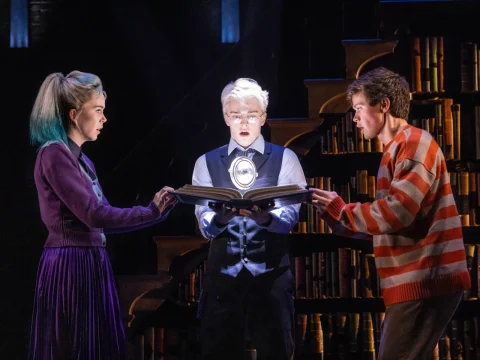 Three actors on stage hold an open, glowing book in front of a bookshelf. The actor in the center wears glasses and a vest, flanked by two others in colorful outfits, appearing surprised.