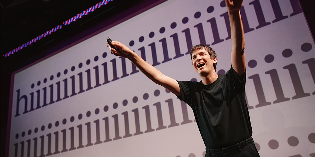 A person in a black shirt stands on stage with arms raised in front of a screen displaying the word "hiiiiiiiiiiiiiiiiiiiiii.