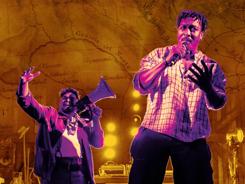 Two performers are on stage. One raises a megaphone while the other speaks into a microphone. They are set against a backdrop of a historical map.