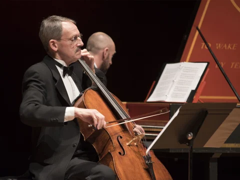 A cellist and a harpsichordist perform on stage. The cellist, seated and dressed in a tuxedo, plays with focus. The harpsichordist, partially visible, has sheet music open in front of him.