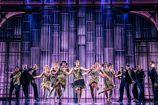 A group of performers in 1920s-style costumes dance in unison on stage with a purple-lit backdrop featuring vertical metallic bars.