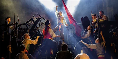 Actors in historical costumes stand on a barricade set, with one person raising a fist under dramatic stage lighting, during a theatrical performance.