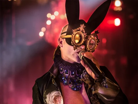 A performer in an ornate rabbit mask and decorative costume sings into a microphone on a dramatically lit stage.
