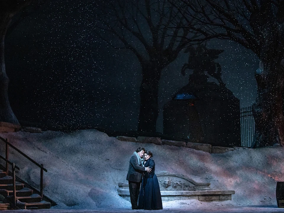 Two people stand embracing in the center of a snowy, nighttime outdoor scene with bare trees and a statue in the background.