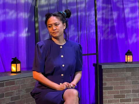 A person with two buns in their hair sits on a brick structure, under purple lighting, and next to small lanterns. They are wearing a dark blue dress with gold buttons and have a thoughtful expression.