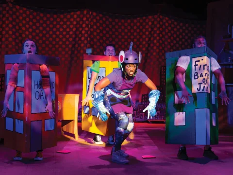 Four performers on stage dressed in colorful cardboard costumes resembling buildings, with one central performer wearing a metallic helmet and gloves amidst a backdrop of red polka dots.